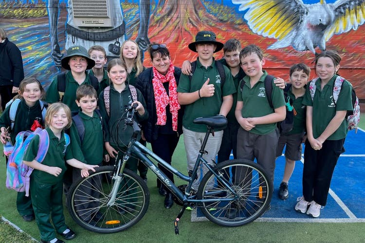 a picture of a group of students standing around thier teacher in front of a colourful mural with a pushbike.
